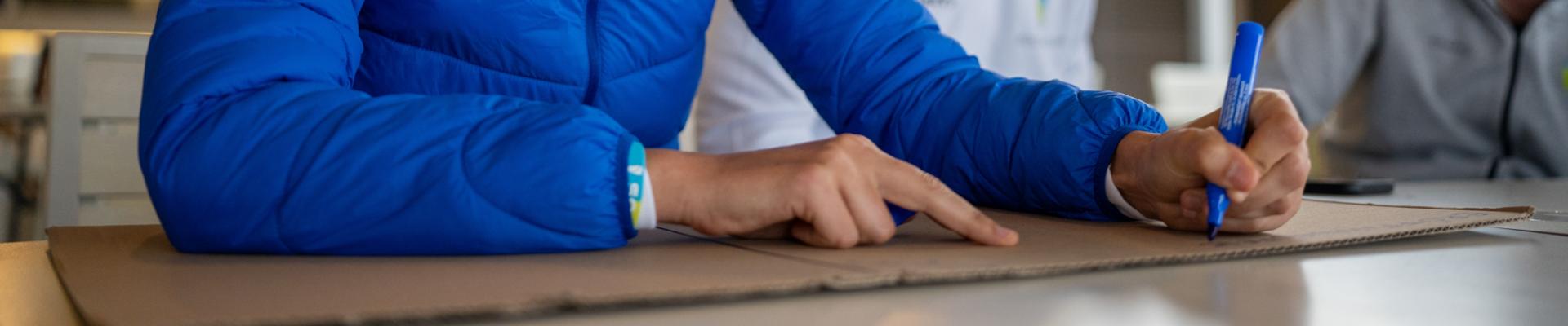 Person in blauer Jacke schreibt mit einem blauen Marker auf einen Karton, während eine andere Person daneben sitzt.