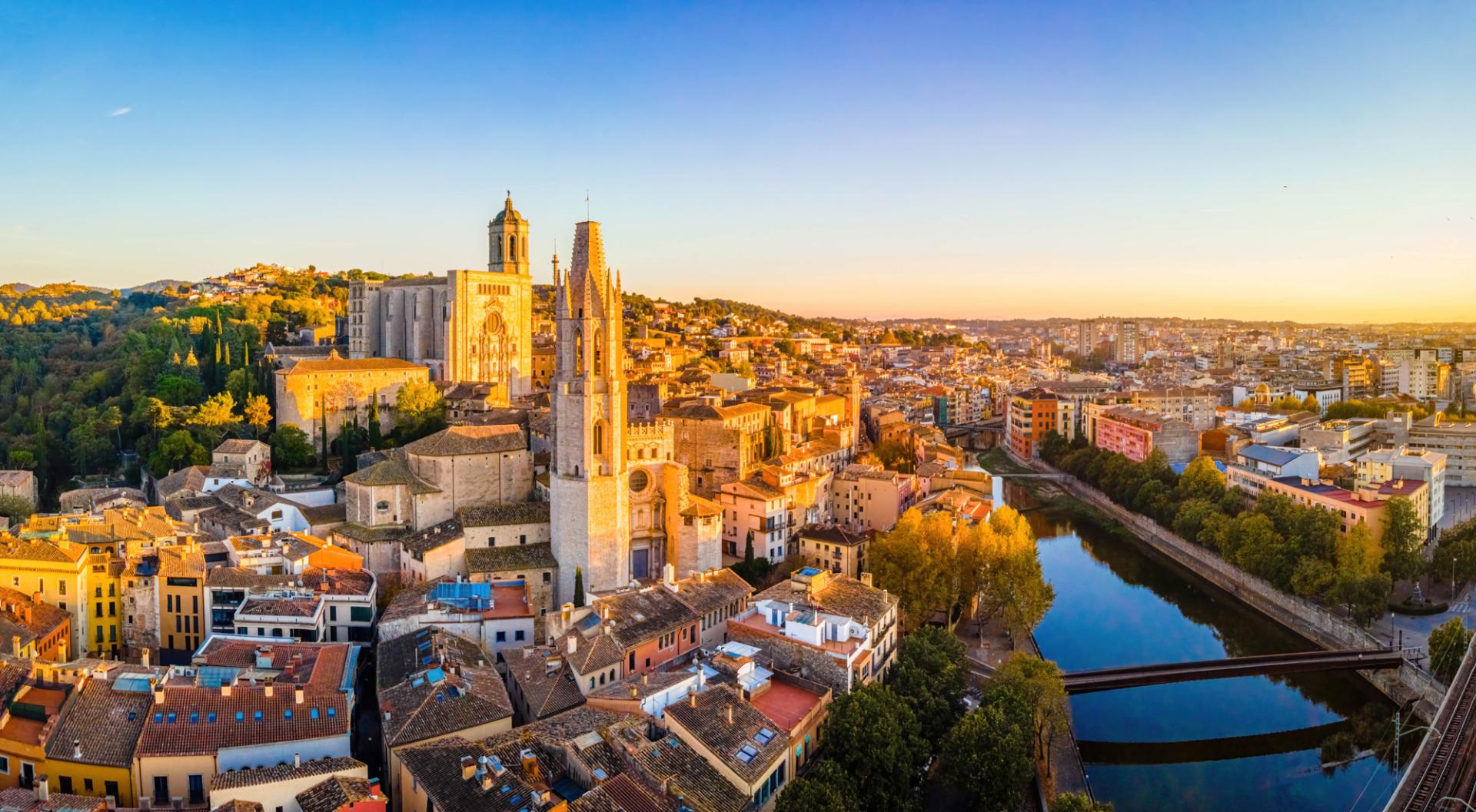 Die Altstadt von Girona in Spanien bei Sonnenuntergang, mit beeindruckenden gotischen und romanischen Bauwerken, umgeben von modernen Stadtteilen und einem ruhigen Fluss, der durch die Stadt fließt