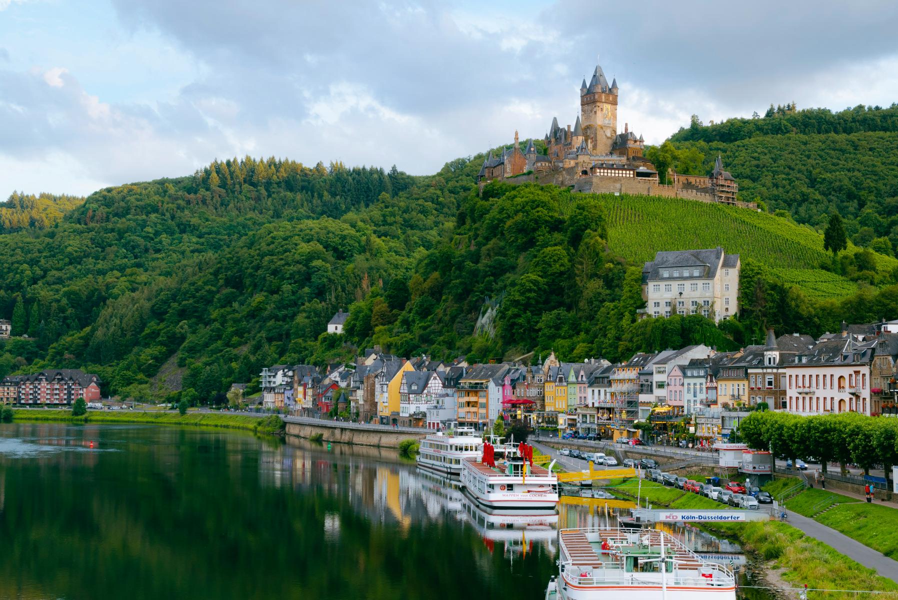 Historische Burg auf einem bewaldeten Hügel, darunter eine Stadt mit bunten Häusern und Schiffen am Flussufer