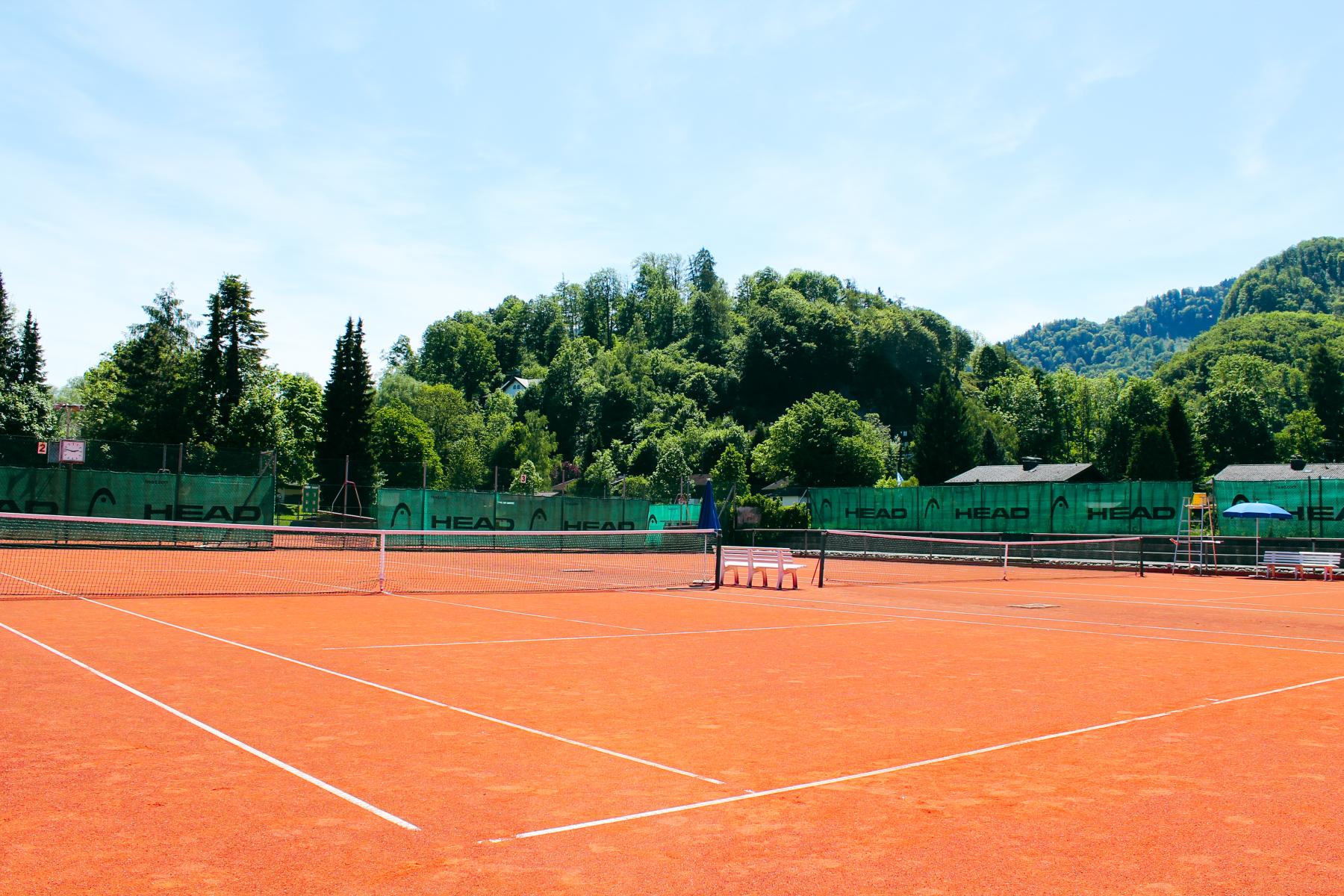 Roter Sand-Tennisplatz in malerischer Umgebung mit grünen Hügeln und Wäldern im Hintergrund, umgeben von grünen Werbebannern.