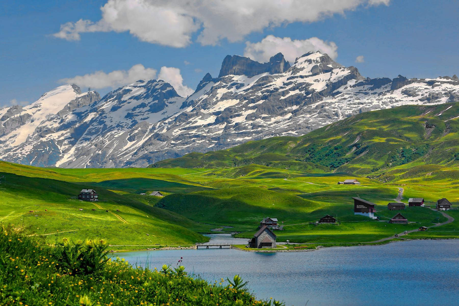 Alpenlandschaft mit See, grünen Wiesen, Holzhäusern und schneebedeckten Bergen im Hintergrund