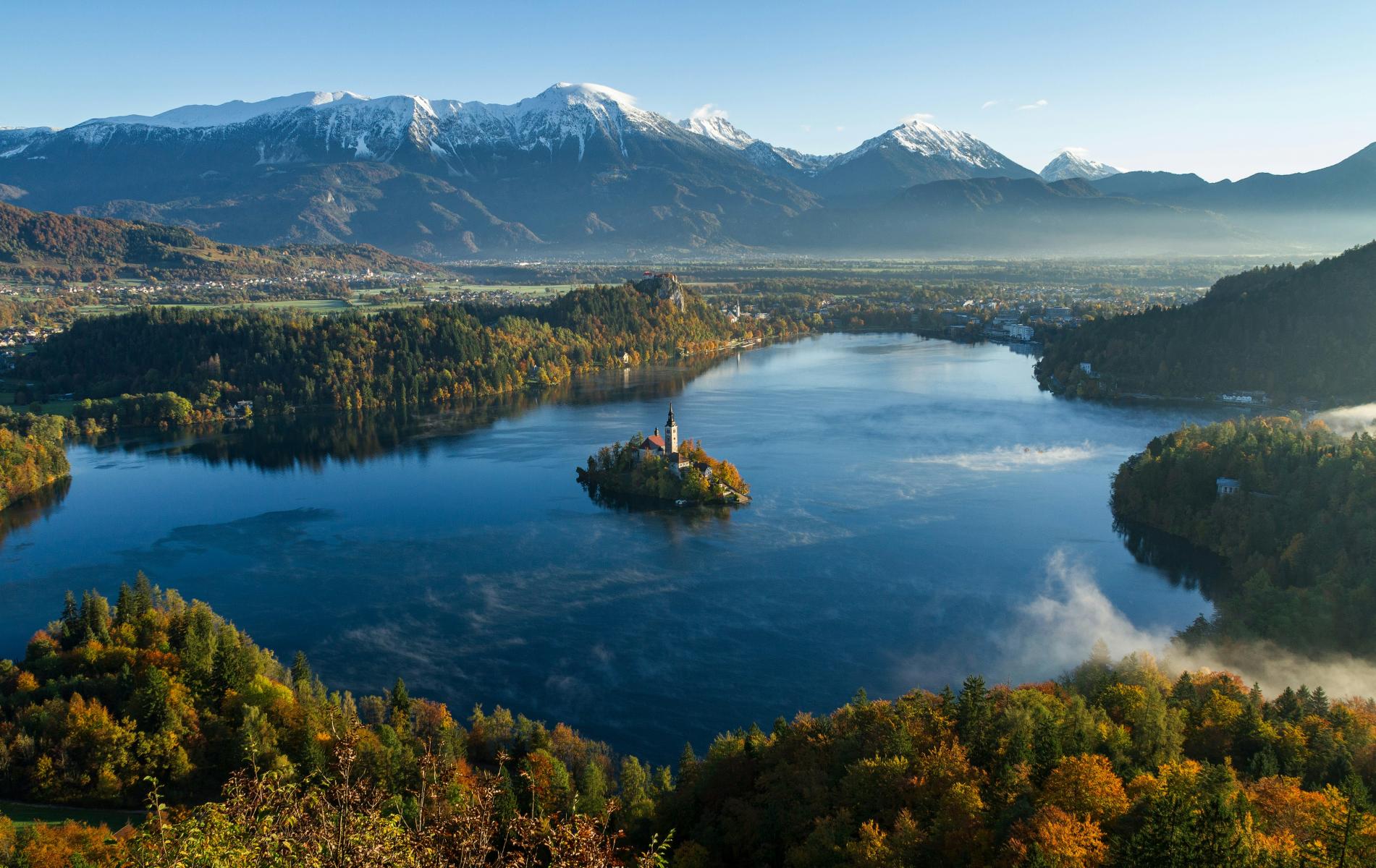 Der malerische Bleder See in Slowenien mit einer Insel in der Mitte, umgeben von herbstlich gefärbten Wäldern und imposanten Bergen im Hintergrund