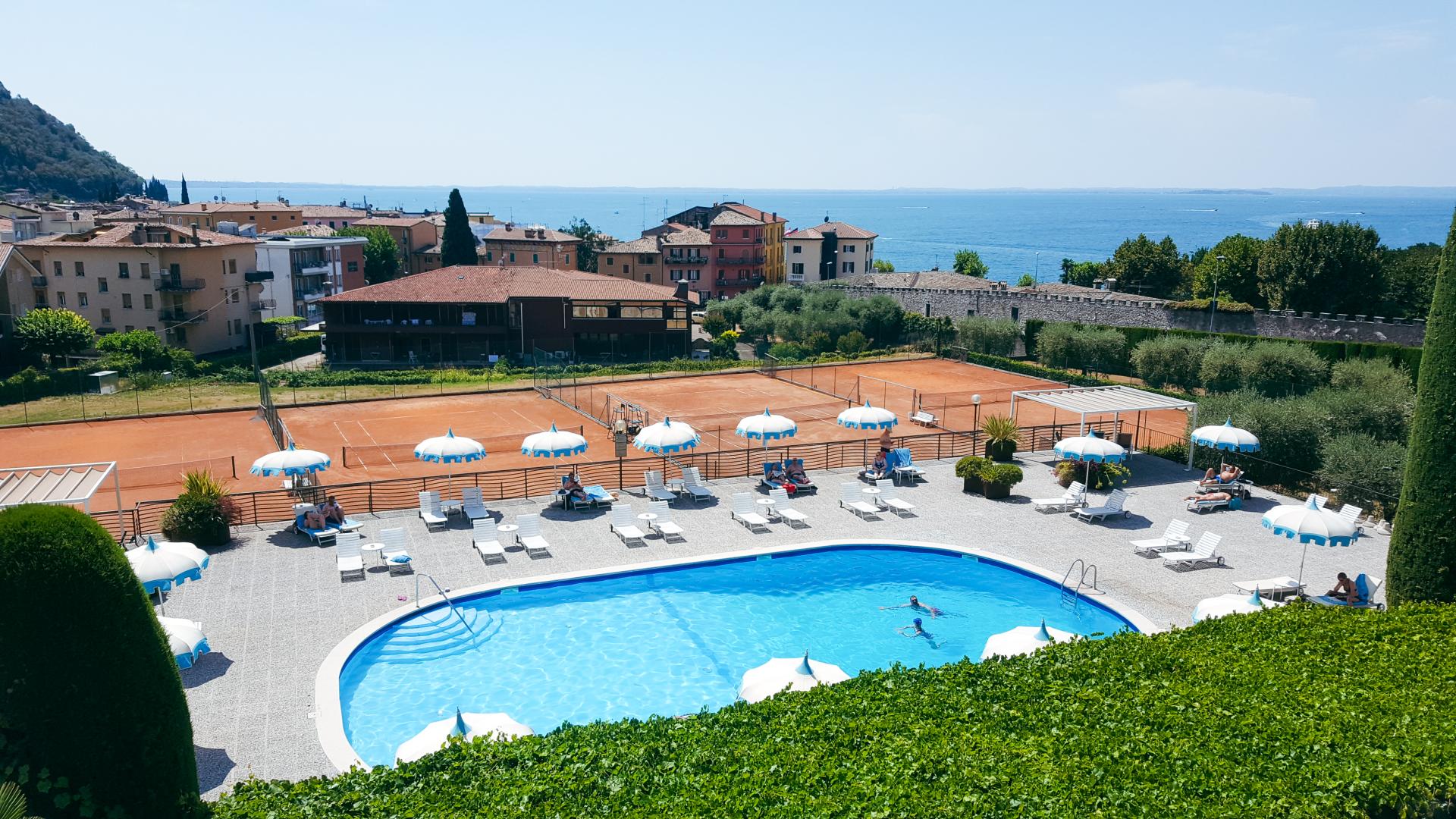 Hotel mit Pool und angrenzenden Tennisplätzen in einer mediterranen Umgebung mit Blick auf einen See.