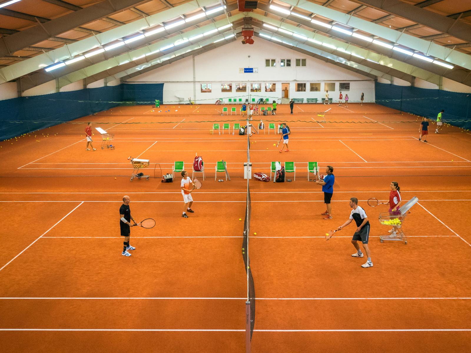 Eine moderne Tennishalle mit mehreren Sandplätzen, auf denen Spieler trainieren und Trainer Übungen anleiten.