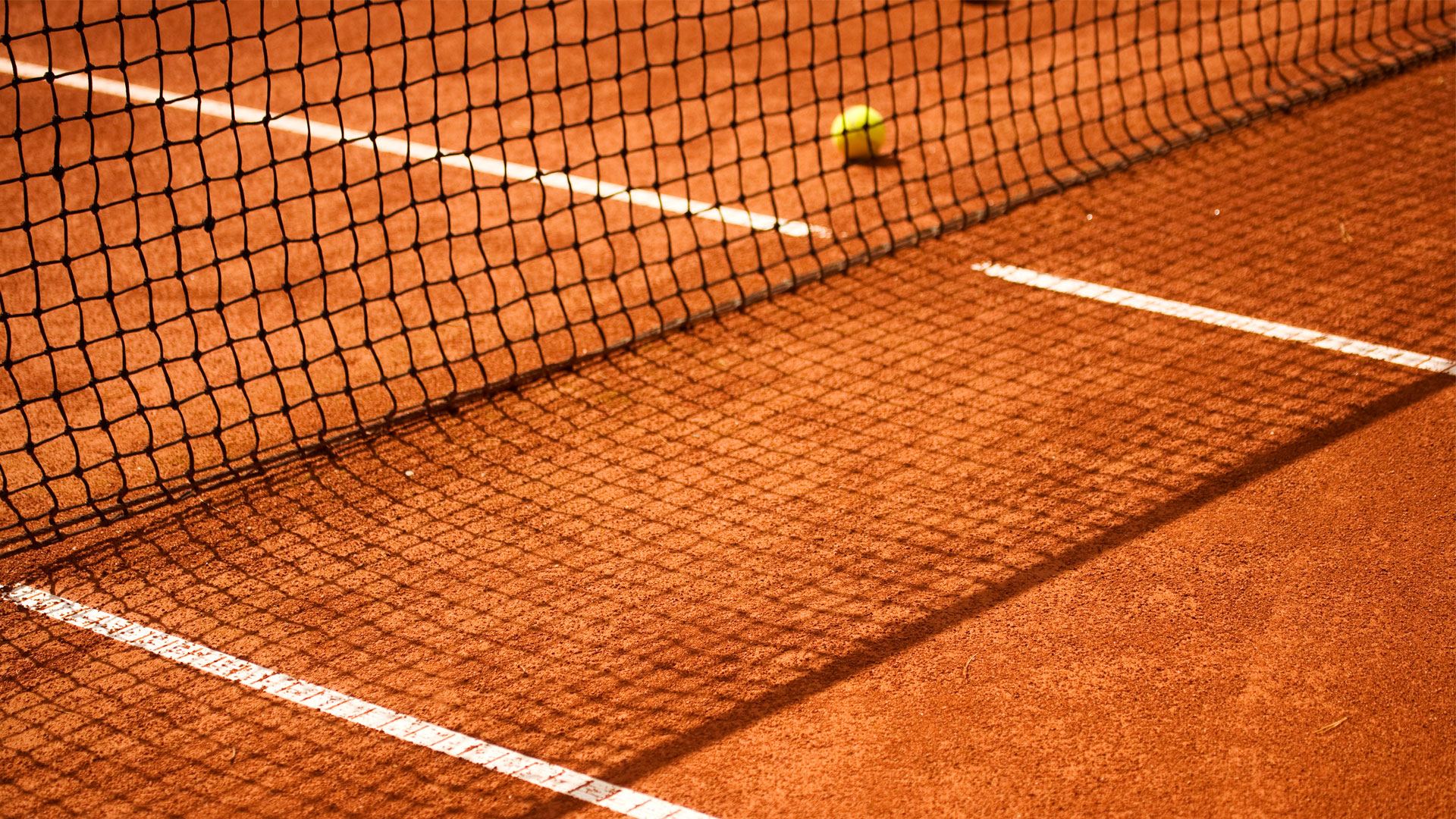 Nahaufnahme eines Sandplatz-Tennisnetzes mit einem gelben Tennisball im Hintergrund, das Spielgeschehen andeutend.