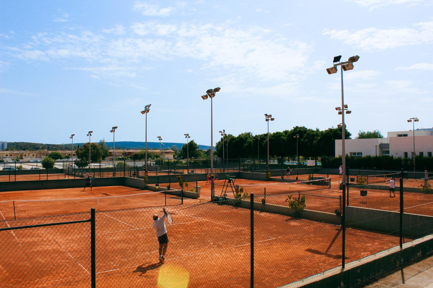 Tennisplätze unter freiem Himmel mit rotem Sandbelag, umgeben von Flutlichtmasten und einer mediterranen Landschaft.