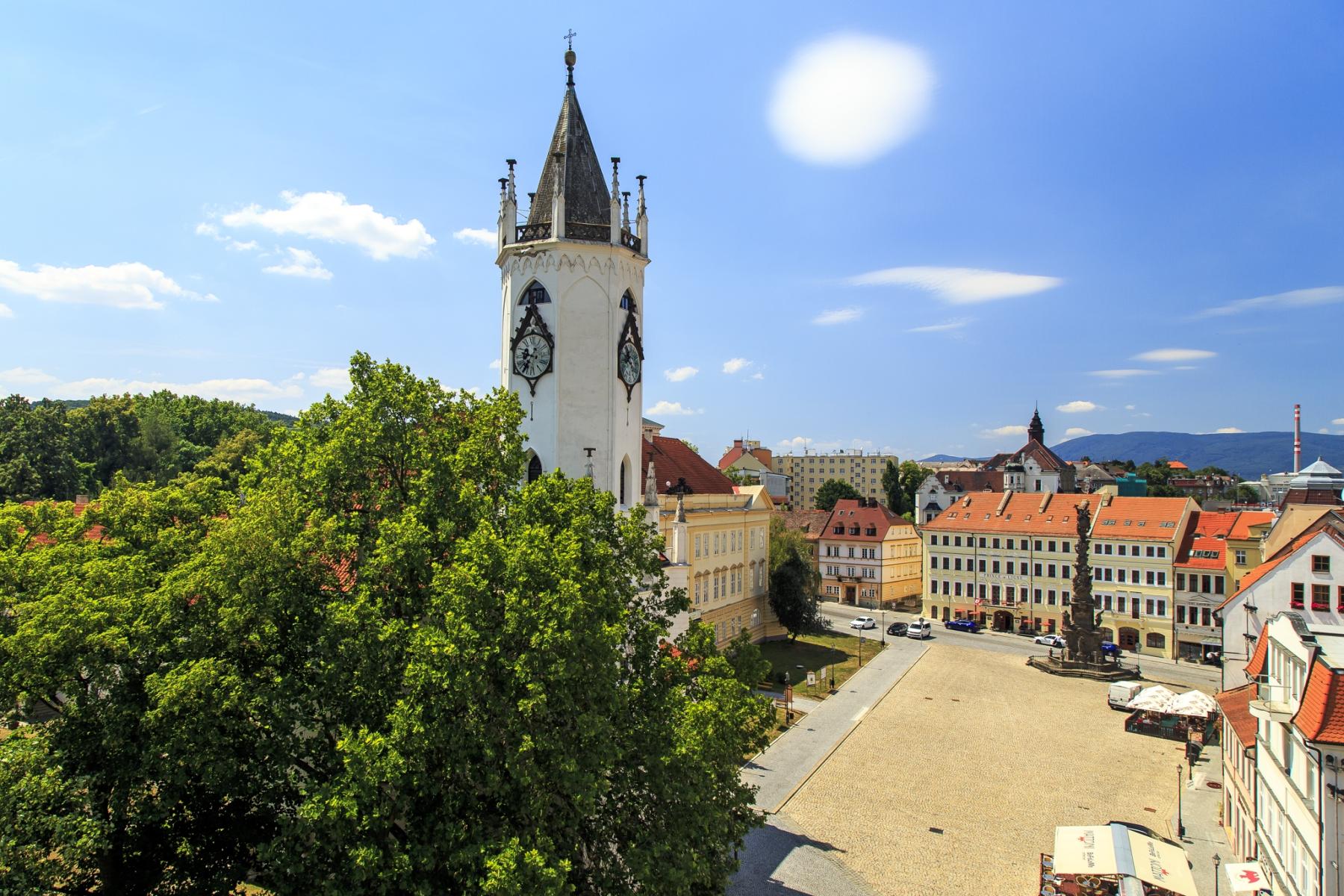 Historischer Marktplatz in Tschechien mit einem markanten Uhrturm im Vordergrund, umgeben von klassischen Gebäuden und einer gepflasterten Fläche unter einem strahlend blauen Himmel