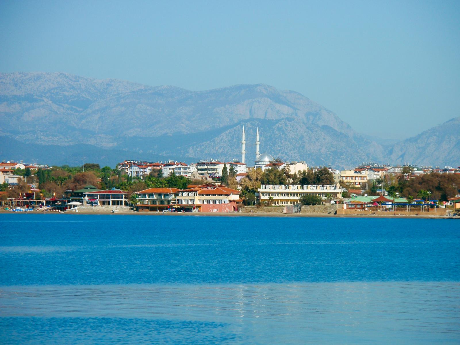 Küstenstadt in der Türkei mit einer Moschee und zwei Minaretten, umgeben von Gebäuden, vor einer beeindruckenden Bergkulisse und dem strahlend blauen Meer im Vordergrund