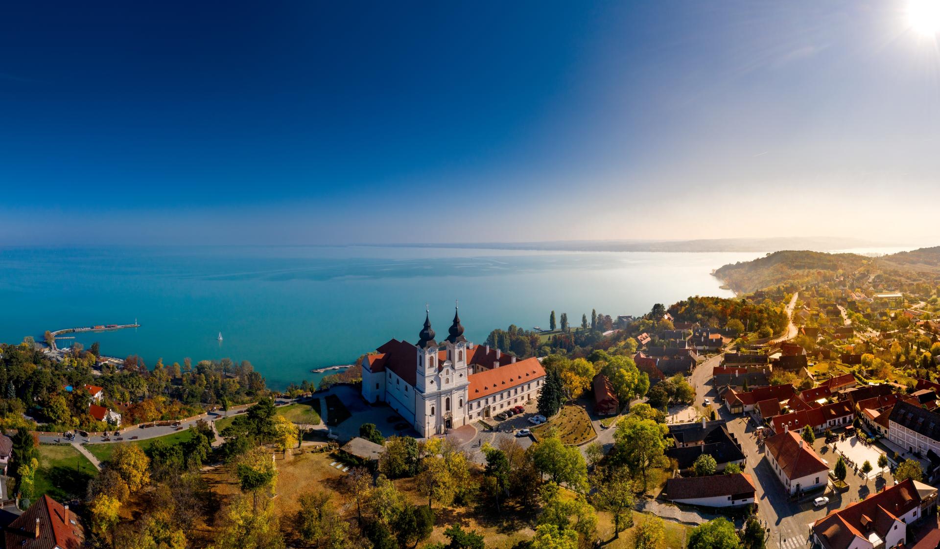 Luftaufnahme eines Klosters mit roten Dächern und zwei Kirchtürmen, umgeben von einer malerischen Landschaft mit Bäumen und Häusern, direkt am Balaton-See unter einem strahlend blauen Himmel