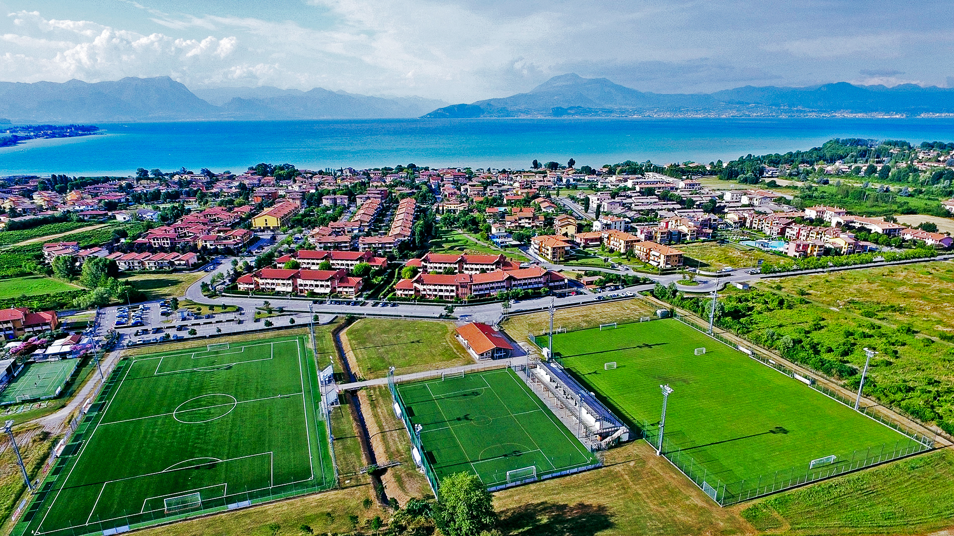 Fußballplätze inmitten einer mediterranen Stadtlandschaft mit Blick auf einen blauen See und umliegende Berge.