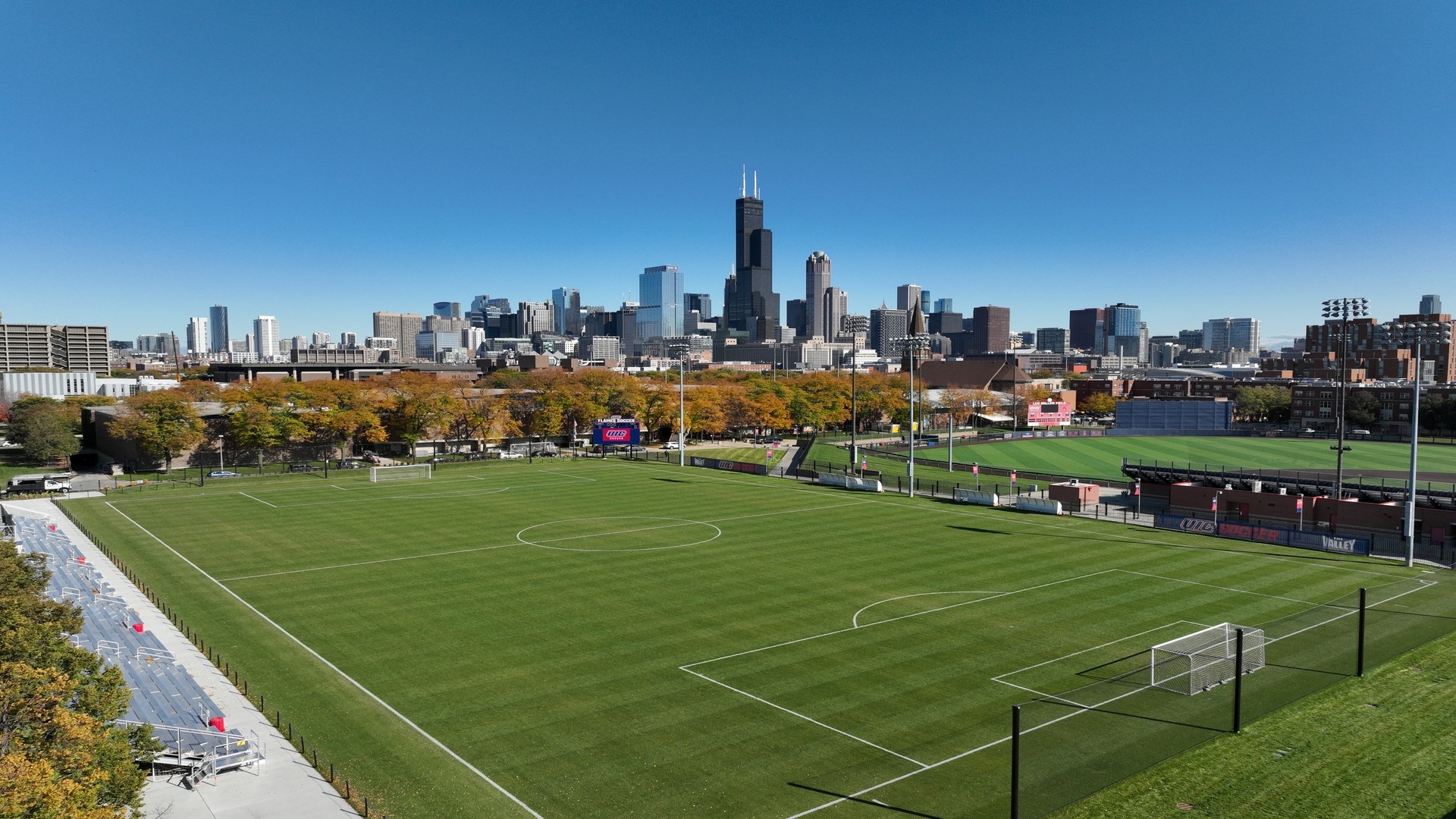 Fußballfeld mit gepflegtem Rasen vor einer modernen Skyline, umgeben von Flutlichtmasten und Tribünen unter klarem, blauem Himmel.