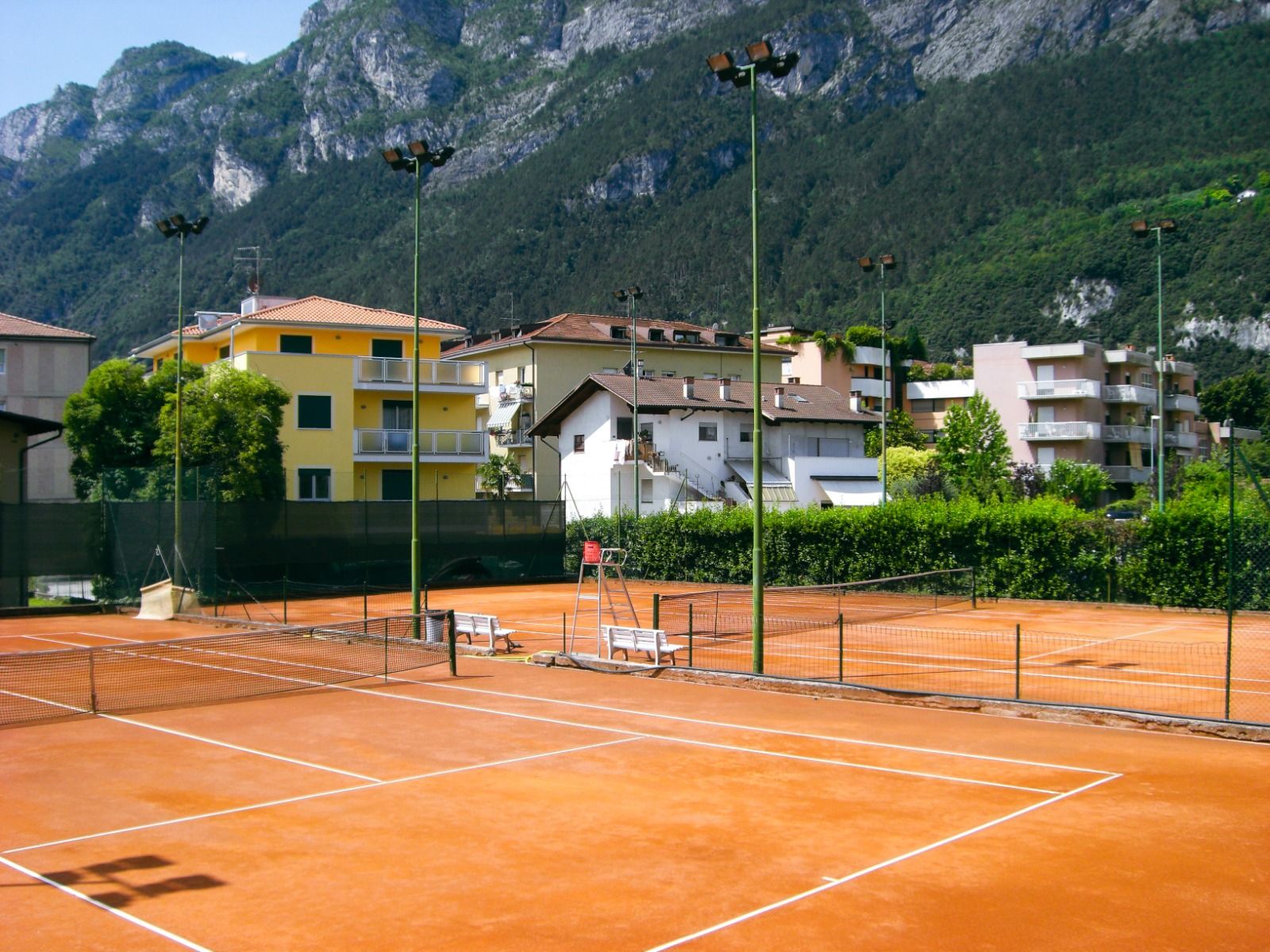 Tennisplatz auf Sand mit Flutlichtanlagen und umliegenden Wohnhäusern in den Bergen.